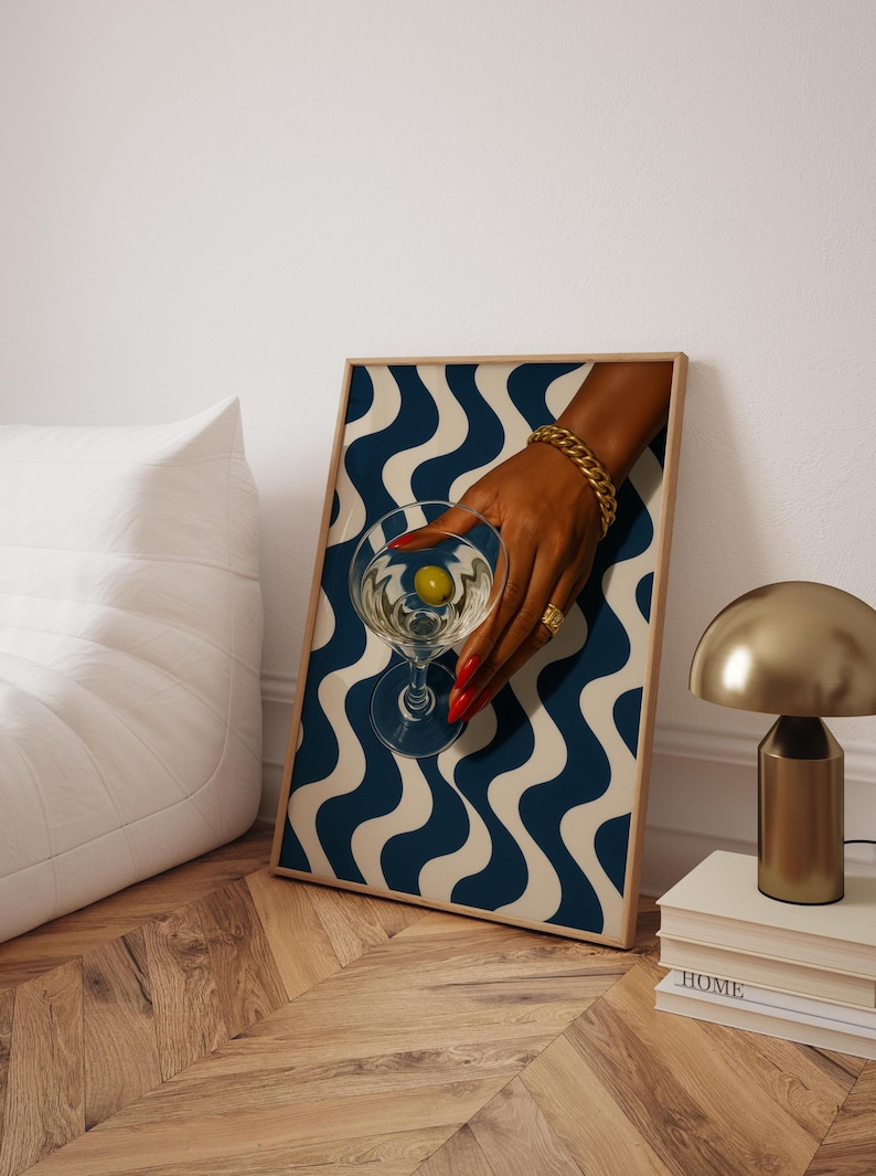 Peut inclure: Une affiche encadr&eacute;e repr&eacute;sentant une main tenant un verre &agrave; martini avec une olive. Le fond pr&eacute;sente un motif ondul&eacute; bleu et blanc. La main a du vernis &agrave; ongles rouge, un bracelet en or et une bague. Une lampe dor&eacute;e et des livres sont en arri&egrave;re-plan.