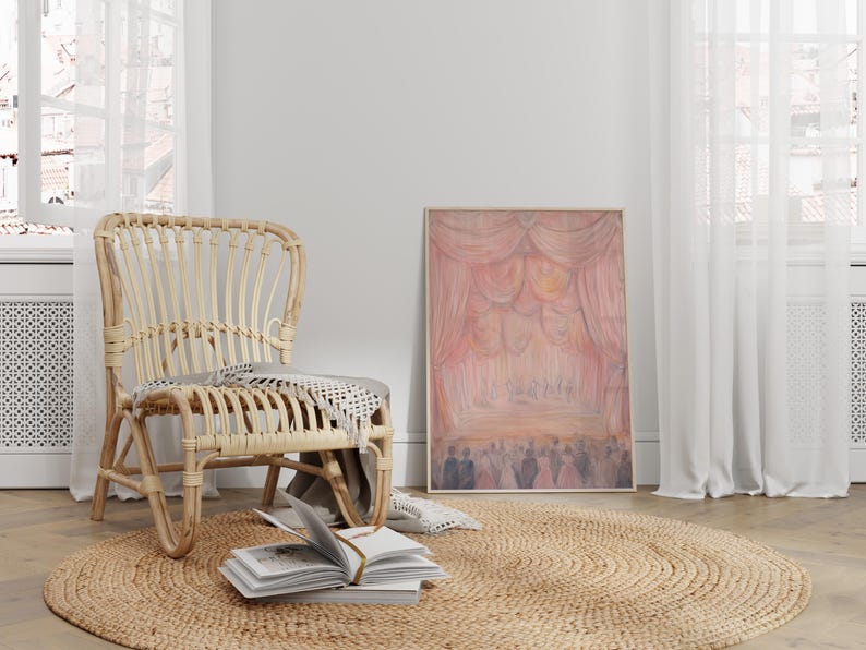May include: A light brown wicker chair with a woven seat and a throw blanket draped over the back. The chair is sitting on a light brown oval rug in a room with white walls and white curtains. A framed painting of a pink stage curtain with people in the background hangs on the wall.