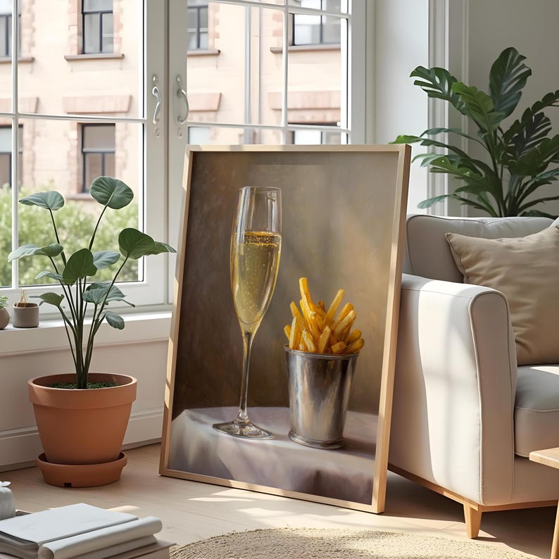 May include: A framed artwork depicts a champagne flute and a metal bucket filled with french fries. The champagne glass is tall and slender, filled with bubbly liquid. The fries are golden brown. The artwork is leaning against a wall near a window.