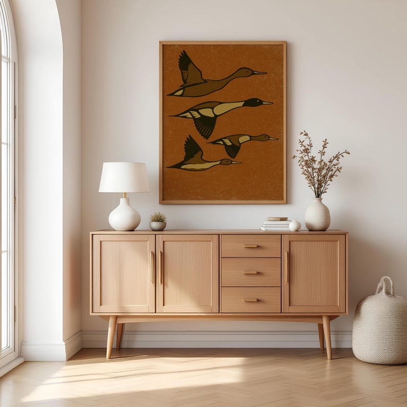 Pu&ograve; includere: Una stampa artistica incorniciata con quattro anatre stilizzate in volo. L'opera d'arte ha uno sfondo marrone caldo con le anatre raffigurate in tonalit&agrave; di marrone e beige. La stampa &egrave; esposta sopra un mobile in legno.