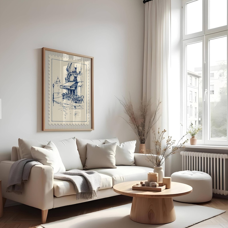 May include: A framed print depicting a blue and white cafe scene, designed to look like a postage stamp. The artwork is above a white sofa with cushions and a grey throw. A wooden coffee table and a white footstool are in front of the sofa.