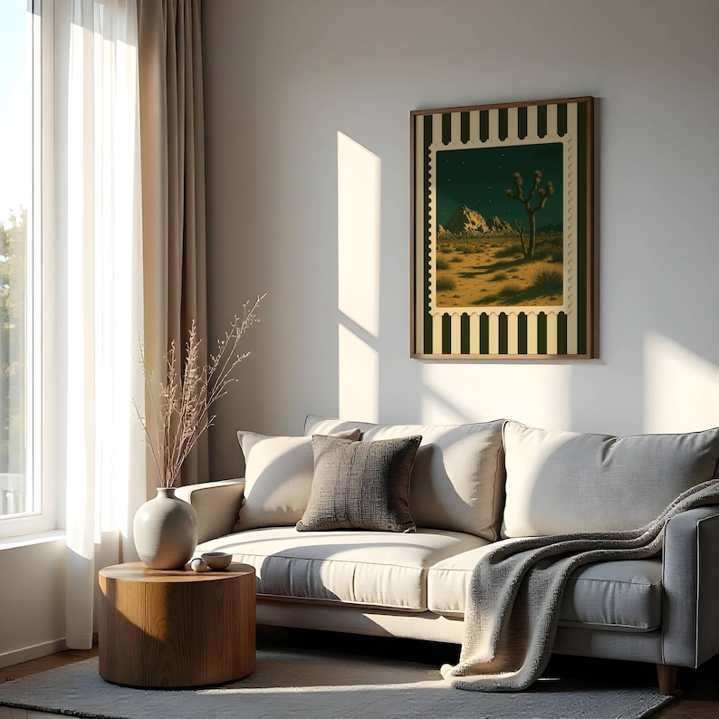 May include: A framed art print featuring a desert landscape with a Joshua tree under a starry night sky. The print has a postage stamp design with a dark green and cream border. The artwork hangs above a cream-colored sofa.