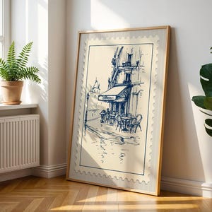 May include: A framed print depicting a cafe scene in blue ink on a cream background. The artwork is in a light wood frame and leans against a white wall. A fern in a terracotta pot sits on a windowsill.