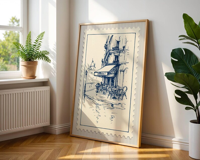 May include: A framed print depicting a cafe scene in blue ink on a cream background. The artwork is in a light wood frame and leans against a white wall. A fern in a terracotta pot sits on a windowsill.