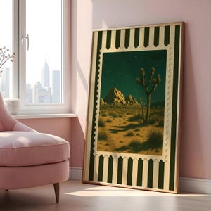 May include: A framed art print featuring a desert landscape with Joshua trees under a starry night sky. The artwork is framed with a green and cream striped border and a gold-colored frame. The print is leaning against a pink wall.