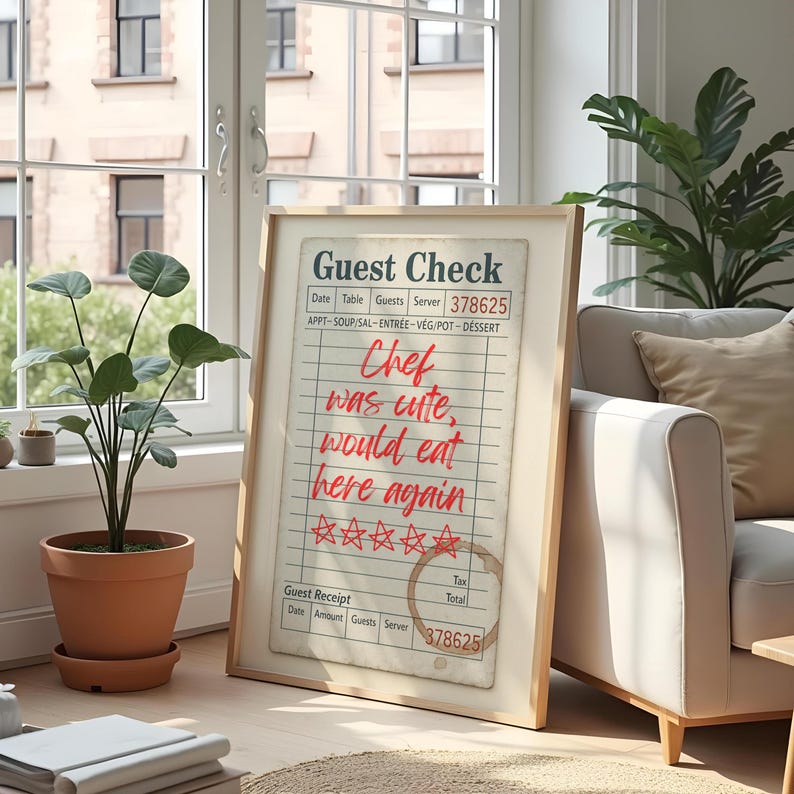 May include: A framed guest check print with a wooden frame. The check has the words "Guest Check" at the top, with the text "Chef was cute, would eat here again" in red. The print is displayed in a well-lit room with plants.