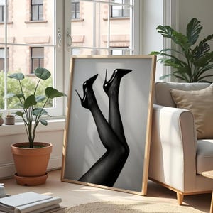 May include: A framed black and white art print featuring a pair of legs in black sheer stockings and high-heeled shoes. The stiletto heels are glossy and pointed, with the legs angled upwards. The artwork is displayed in a light-colored wooden frame.
