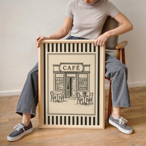 May include: A framed black and white illustration of a cafe with the word "CAF&Eacute;" above the entrance. The artwork features tables, chairs, and floral arrangements. The frame is natural wood with black and white striped borders.