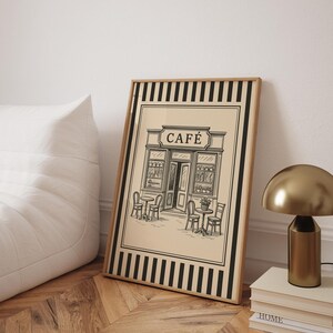 May include: Framed art print featuring a black and white illustration of a cafe with outdoor seating. The word "CAF&Eacute;" is displayed above the entrance. The print has a beige background with a black and white striped border and a wooden frame.