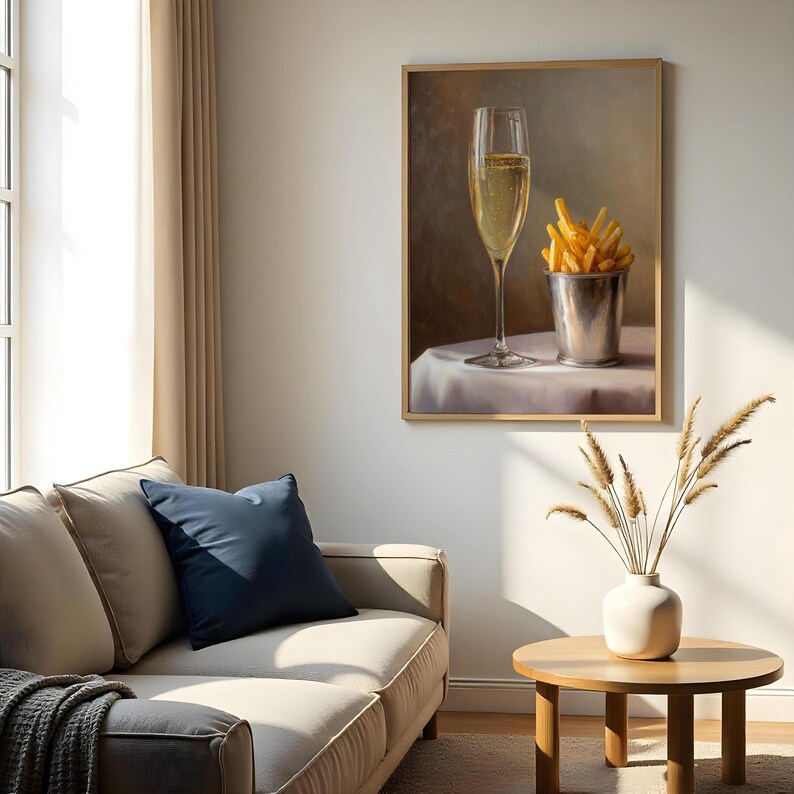 May include: A framed painting depicts a champagne flute and a metal bucket filled with french fries on a white tablecloth. The painting is in a room with a beige sofa, a blue pillow, and a wooden side table.