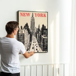 May include: A person hangs a black framed poster on a white wall. The poster features a black and white illustration of the New York City skyline with the words "NEW YORK" in red letters at the top.