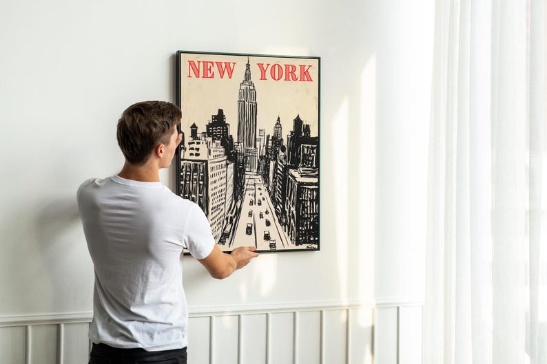 May include: A person hangs a black framed poster on a white wall. The poster features a black and white illustration of the New York City skyline with the words "NEW YORK" in red letters at the top.