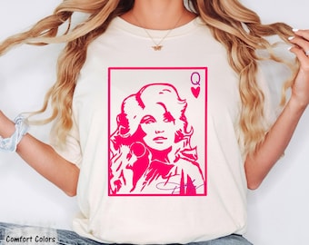 May include: An ivory short-sleeve t-shirt featuring a hot pink graphic print of a woman with voluminous hair styled like a playing card. The design includes the letter Q and a heart symbol in the top right corner, with a signature at the bottom.