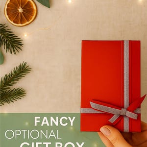 A hand holding a red rectangular gift box with a silver ribbon bow, placed on a beige background with pine leaves, dried orange slices, and warm fairy lights. The text reads “Fancy Optional Gift Box.”
