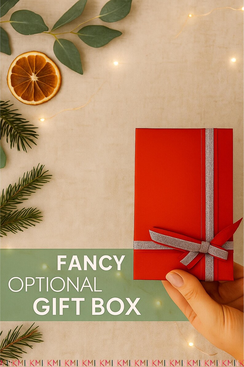 A hand holding a red rectangular gift box with a silver ribbon bow, placed on a beige background with pine leaves, dried orange slices, and warm fairy lights. The text reads “Fancy Optional Gift Box.”