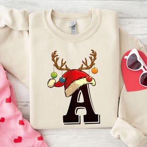 Puede incluir: Sudadera color crema con un diseño navideño. El diseño presenta un gorro de Papá Noel rojo con cuernos de reno, adornos y la letra "A". También se ven unas gafas de sol en forma de corazón y un objeto rojo en forma de corazón.