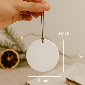 Hand holding a 3-inch round ceramic ornament with a green cord hanger on a neutral boho Christmas background. Styled with pine twigs, dried orange slices, linen fabric, and warm fairy lights for a cozy holiday look.