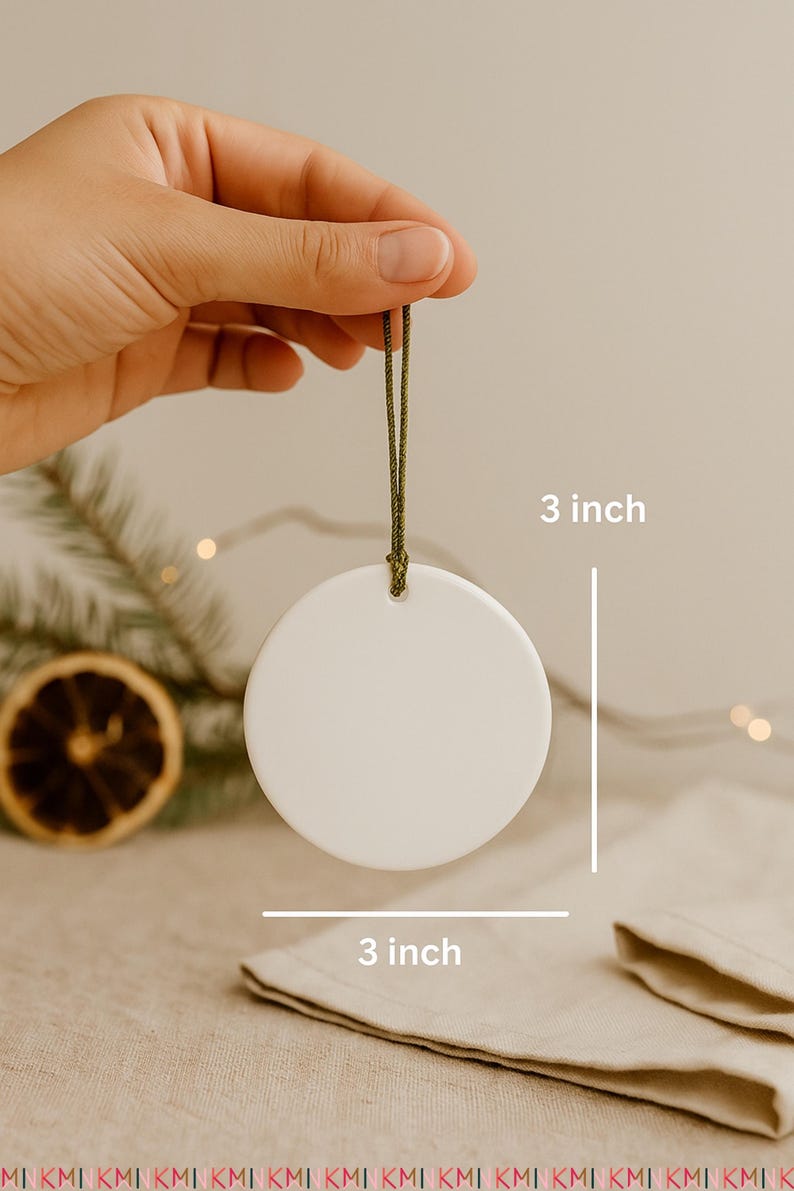 Hand holding a 3-inch round ceramic ornament with a green cord hanger on a neutral boho Christmas background. Styled with pine twigs, dried orange slices, linen fabric, and warm fairy lights for a cozy holiday look.