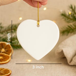 Hand holding a 3-inch heart-shaped ceramic ornament with a gold hanging string on a beige boho Christmas background. Includes pine branches, dried orange slices, and soft fairy lights for scale and festive atmosphere.