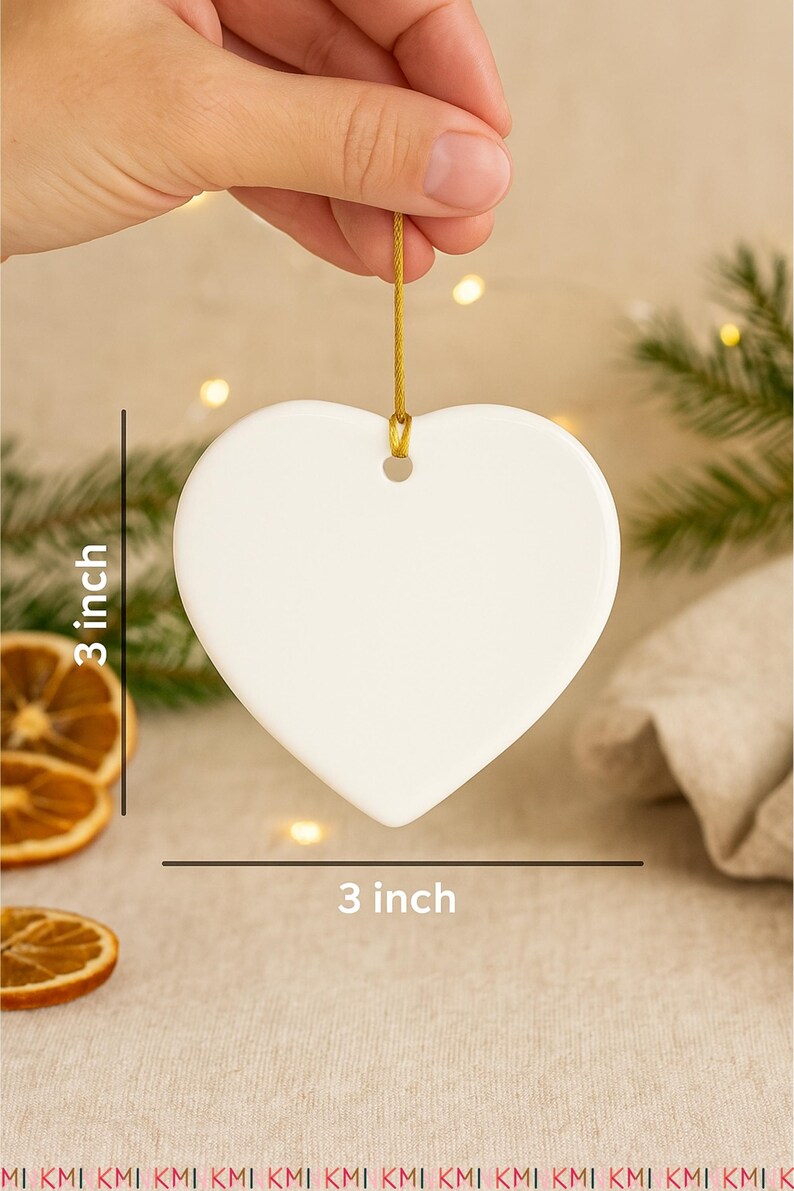 Hand holding a 3-inch heart-shaped ceramic ornament with a gold hanging string on a beige boho Christmas background. Includes pine branches, dried orange slices, and soft fairy lights for scale and festive atmosphere.