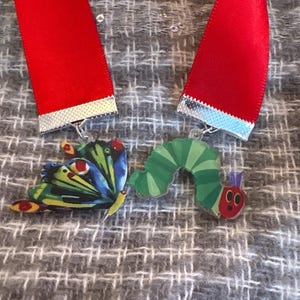 May include: A necklace featuring a colorful butterfly and caterpillar pendant. The pendants are attached to a red ribbon with silver trim. The butterfly has blue, green, and yellow wings. The caterpillar is green with red accents.
