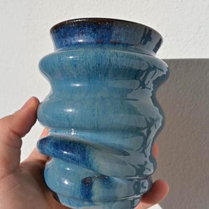 Könnte beinhalten: Eine handgefertigte Keramikvase mit einem einzigartigen, strukturierten Design. Die Vase hat eine hellblaue Glasur mit dunkelblauen Akzenten, die ein wirbelndes Muster erzeugt. Der Rand ist ein dunklerer Blauton. Die Vase wird von einer Hand gehalten.