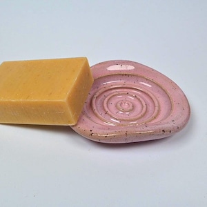 Könnte beinhalten: Ein rechteckiger gelber Seifenblock liegt auf einer rosa Keramik-Seifenschale. Die Schale hat ein Spiralmuster und eine gesprenkelte Oberfläche. Seife und Schale stehen vor einem weißen Hintergrund.
