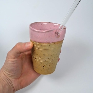 Könnte beinhalten: Eine handgefertigte Keramiktasse mit rosa Rand und einem beigefarbenen, strukturierten Körper. Die Tasse hat einen durchsichtigen Glasstrohhalm eingesetzt. Die Tasse wird in einer Hand vor weißem Hintergrund gehalten. Die Tasse ist wahrscheinlich zum Trinken.
