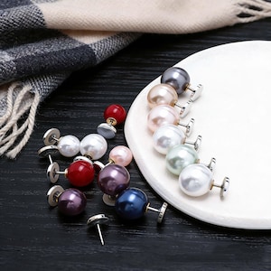 Op de afbeelding: Een verzameling parelstud-oorbellen in verschillende kleuren, waaronder wit, roze, rood, paars en blauw. De oorbellen worden gepresenteerd op een wit bord en een zwart oppervlak, met een sjaal op de achtergrond.