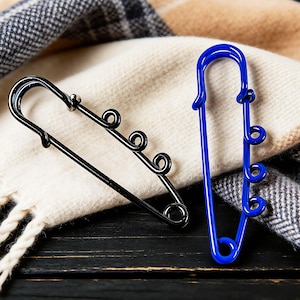 May include: Two decorative safety pins, one black and one blue, rest on a cream-colored scarf with fringed edges. The pins have a unique design with decorative swirls. The background includes a plaid pattern and a dark wooden surface.