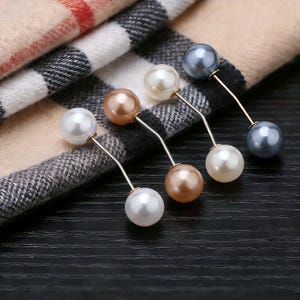 May include: A collection of pearl pins in various colors, including white, bronze, and gray, with gold-colored stems. The pins are arranged on a plaid scarf and a dark wooden surface, showcasing their elegant design.