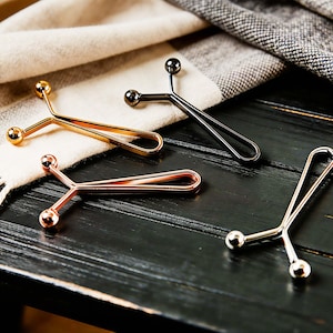 May include: Four metal scarf clips in gold, silver, rose gold, and black. Each clip has a Y-shaped design with a small ball at each end. The clips are displayed on a dark wooden surface with a cream and grey scarf.