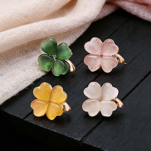 May include: Four clover-shaped brooches in green, pink, yellow, and white. Each brooch has four heart-shaped petals and a gold-colored stem. The brooches are displayed on a black surface with a light pink fabric in the background.
