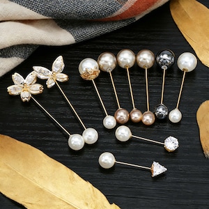 May include: A collection of decorative pins with various designs. The pins feature pearls, crystals, and floral or geometric shapes. The pins have gold-colored stems and are arranged on a dark surface.