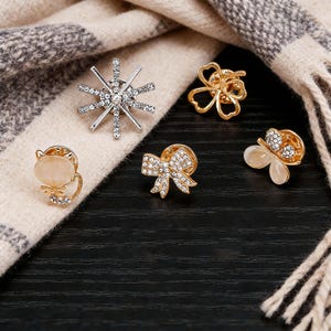 May include: An assortment of decorative pins, including a snowflake, a four-leaf clover, a bow, a cat, and a butterfly. The pins are gold and silver-toned, with rhinestone accents. The pins are displayed on a black surface with a beige and gray scarf.