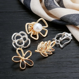Puede incluir: Una colección de broches decorativos con diseños florales y de hojas. Los broches son en tonos dorados y plateados, uno de ellos con un centro de perla. Están dispuestos sobre una superficie de madera oscura con una tela estampada en el fondo.
