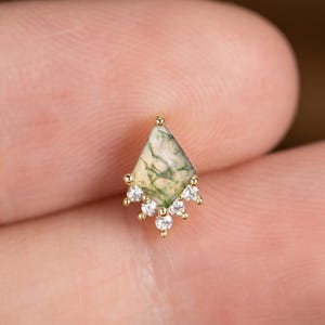 May include: A small, gold-colored earring with a diamond-shaped, green and white stone. The earring is adorned with small, clear gemstones at the bottom, creating a delicate, elegant design. The earring is shown on a finger.