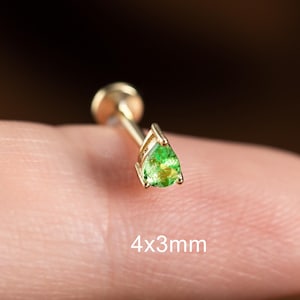 May include: A gold earring featuring a pear-shaped, green gemstone. The gemstone is set in a fine frame. The text "4x3mm" is printed below the earring, indicating its size. The earring is displayed against a blurred background.