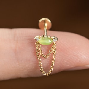 May include: A gold-coloured earring featuring a light green oval-shaped stone and two small clear stones. Two delicate gold chains hang beneath the stone. The earring is designed for a piercing.