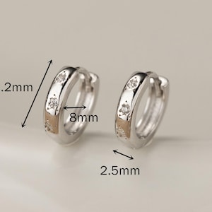 May include: A pair of silver-colored hoop earrings with small, clear gemstones. Each earring is 10.2mm long, 8mm wide, and 2.5mm thick. The earrings are displayed on a white surface.