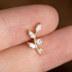 May include: A gold-colored earring in the shape of a delicate leaf branch. The branch features several clear, sparkling cubic zirconia stones. The earring is designed to be worn in a piercing.