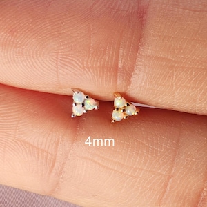 Può includere: Due piccoli orecchini triangolari con un gruppo di pietre di opale bianche. Gli orecchini sono incastonati in metallo color oro. Il testo "4mm" è visibile, indicando la dimensione degli orecchini.