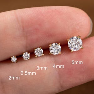 May include: A collection of six gold stud earrings with clear, round cubic zirconia stones. The sizes are labeled as 2mm, 2.5mm, 3mm, 4mm, and 5mm. The earrings are displayed on a person's finger.