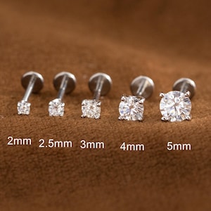 May include: Five silver stud earrings with clear, round gemstones of varying sizes. The sizes are labeled as 2mm, 2.5mm, 3mm, 4mm, and 5mm. The earrings are displayed on a brown surface.