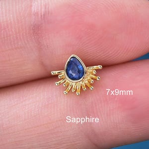 May include: A gold-colored earring with a teardrop-shaped blue sapphire gemstone, measuring 7x9mm. The sapphire is surrounded by a sunburst design. The word "Sapphire" is printed below the earring.