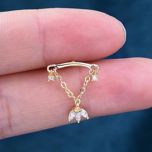 May include: A gold-coloured ear piercing with a curved bar and a dangling chain design. The chain supports three clear, sparkling cubic zirconia stones. The bottom of the chain features a cluster of three stones in a floral arrangement.