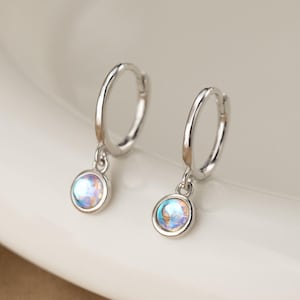 May include: A pair of silver hoop earrings with iridescent, round, dangling charms. The charms have a pearlescent, multi-colored effect, reflecting hues of blue, green, and purple. The earrings are displayed on a white surface.