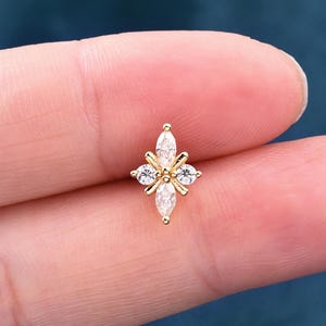 May include: A gold-colored stud earring with a four-petal design. Each petal features a marquise-cut, clear crystal, with smaller round crystals between them. The earring is held between two fingers.