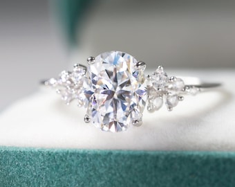 May include: A silver ring with a large, oval-cut, clear gemstone as the focal point. Smaller, round gemstones flank the main stone on each side. The ring is set against a soft, white background inside a teal box.
