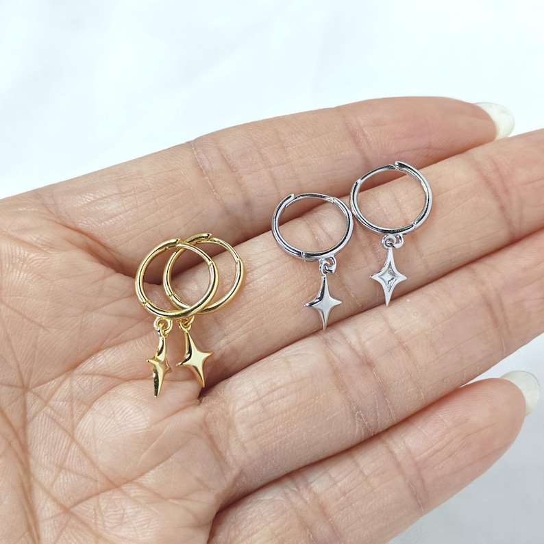 Puede incluir: Dos pares de pendientes de aro, uno dorado y otro plateado, cada uno con un dije de estrella colgante. Los pendientes se muestran sobre un fondo claro, mostrando su dise&ntilde;o delicado. Una joya sencilla y elegante.